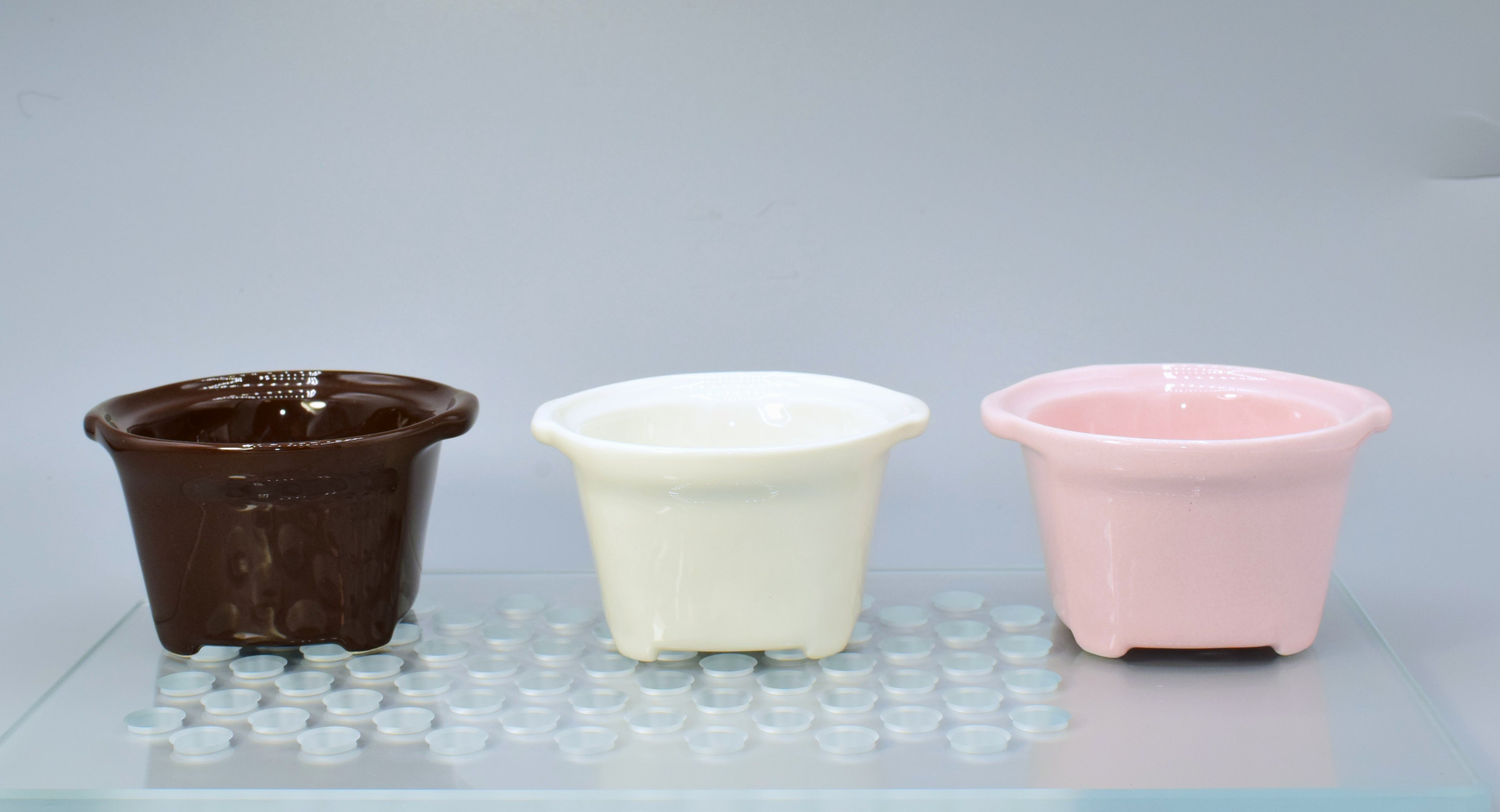 Three ceramic espresso cups in brown, white, and pink on a reflective surface with a light gray background.